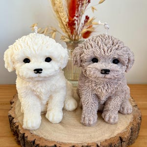 May include: Two realistic-looking dog candles, one white and one grey, are sitting on a wooden slice. The candles are detailed with fur and have black eyes.