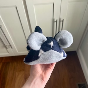 May include: A plush mouse hat in navy blue and light gray. The hat features large, rounded ears and a pointed top, with a patchwork design. The hat is held in a hand.