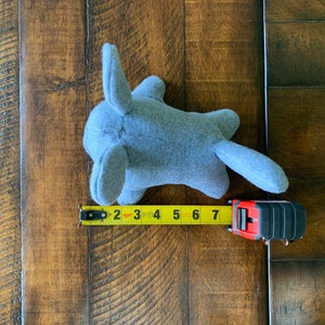 May include: A gray plush toy shaped like a small animal with large ears and a tail. The toy is measured with a yellow tape measure, showing it is 7 inches long.