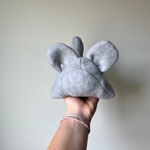 May include: Gray plush toy shaped like a mouse with large ears. The toy is held in a hand with bracelets.