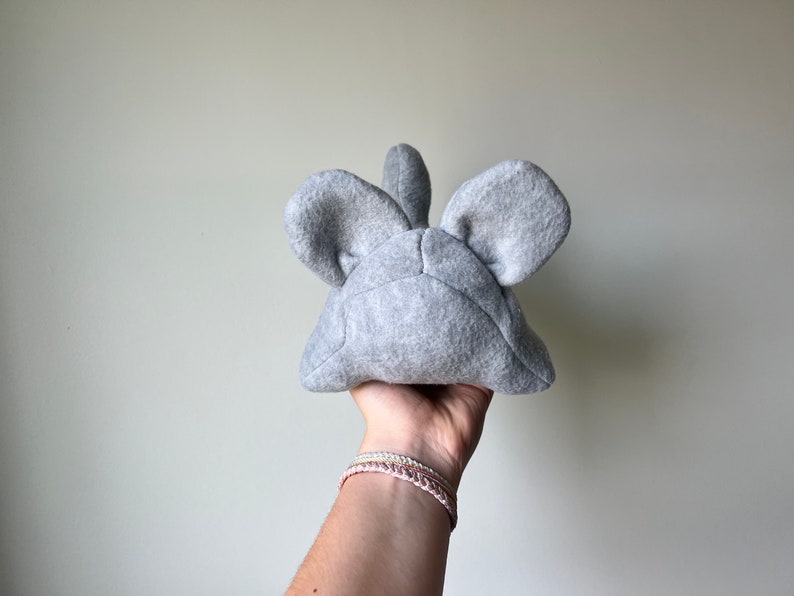 May include: Gray plush toy shaped like a mouse with large ears. The toy is held in a hand with bracelets.