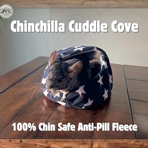 Puede incluir: Un chinchilla asoma de una cueva de abrazo de forro polar azul y blanco con estampado de estrellas. El texto "Chinchilla Cuddle Cove" está encima del producto, y "100% Chin Safe Anti-Pill Fleece" está debajo. El producto está sobre una superficie de madera.