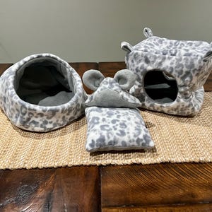 Może przedstawiać: Zestaw akcesoriów dla małych zwierząt domowych, w tym okrągłe legowisko, poduszka w kształcie myszy i domek. Wszystkie elementy wykonane są z miękkiej tkaniny w szaro-biały wzór w cętki. Domek ma okrągły otwór.