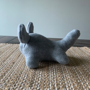 May include: A gray plush toy shaped like a shark with large, floppy ears. The toy is lying on a woven rug.