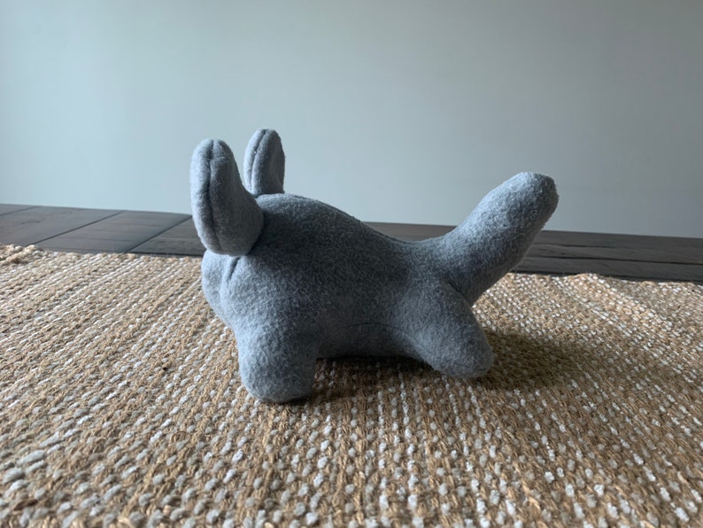 May include: A gray plush toy shaped like a shark with large, floppy ears. The toy is lying on a woven rug.