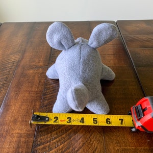 May include: A gray plush toy shaped like a mouse, measuring 7 inches long. The toy is lying on a wooden surface with a measuring tape next to it.