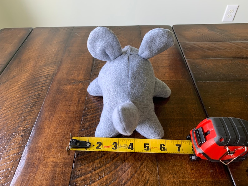 May include: A gray plush toy shaped like a mouse, measuring 7 inches long. The toy is lying on a wooden surface with a measuring tape next to it.