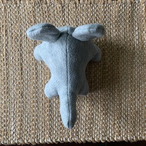 May include: A gray plush toy shaped like an elephant with large ears. The toy is lying on a woven rug.