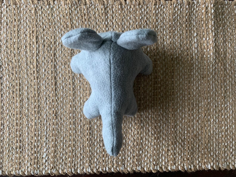 May include: A gray plush toy shaped like an elephant with large ears. The toy is lying on a woven rug.