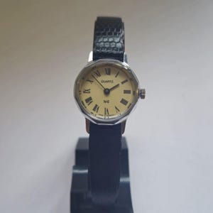 Może przedstawiać: Zegarek kwarcowy w stylu vintage ze srebrną kopertą i kremową tarczą z czarnymi rzymskimi cyframi. Zegarek ma czarny skórzany pasek z teksturowanym wzorem. Zegarek jest prezentowany na czarnym stojaku.
