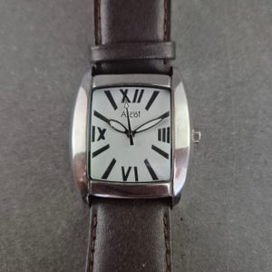 Könnte beinhalten: Rechteckige Armbanduhr mit silberfarbenem Gehäuse und weißem Zifferblatt mit schwarzen römischen Ziffern und Zeigern. Die Uhr hat ein braunes Lederarmband. Der Markenname "Ascot" ist auf dem Zifferblatt aufgedruckt.