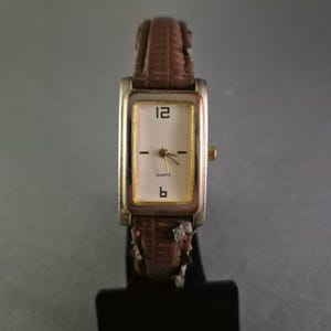 Pode incluir: Um relógio de quartzo retangular com caixa prateada e pulseira de couro castanho. O mostrador é branco com algarismos pretos às 12 e às 6, e traços pretos para os outros marcadores de hora. Os ponteiros são dourados.