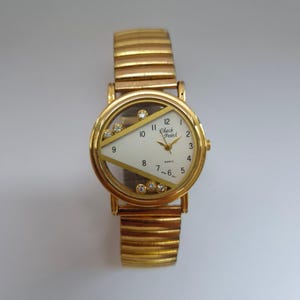 Könnte beinhalten: Goldfarbene Armbanduhr mit weißem Zifferblatt und dem Text "Check Point". Die Uhr hat eine goldene Lünette mit transparenten Akzenten und ein goldfarbenes, dehnbares Armband. Das Zifferblatt hat goldene Zeiger und Stundenmarkierungen.