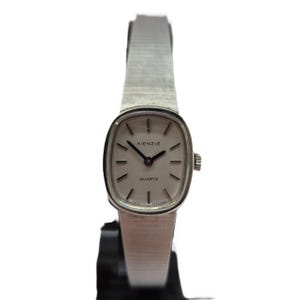 Puede incluir: Un reloj Kienzle Quartz plateado con una esfera rectangular y una correa plateada. La esfera del reloj es de color claro con manecillas e indicadores de hora negros. La palabra "Kienzle" está impresa en la parte superior de la esfera y "QUARTZ" en la parte inferior.