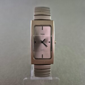 Op de afbeelding: Een rechthoekig TCM quartz horloge met een zilverkleurige metalen band en kast. De wijzerplaat is lichtroze met zwarte wijzers en het woord "TCM" boven de wijzers. Het woord "QUARTZ" staat eronder.