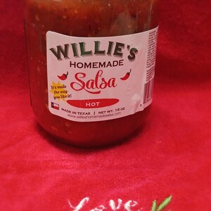 May include: A jar of Willie's Homemade Salsa, labeled "Hot," sits on a red surface embroidered with "Love, Faith, Family." The salsa jar has a gold lid and a white label with red accents. The text "It's made the way you like it!" is also on the label.