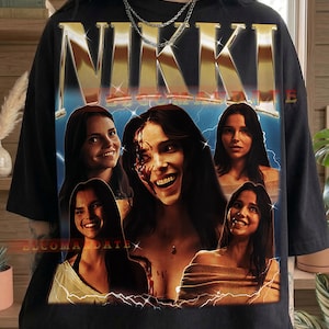 Może przedstawiać: Czarna koszulka z grafiką z napisem "NIKKI" w złotych metalicznych literach nad kolażem zdjęć kobiety. Obrazy są umieszczone na niebiesko-ż&oacute;łtym tle z błyskawicami. Słowo "Black" jest wydrukowane na dole.