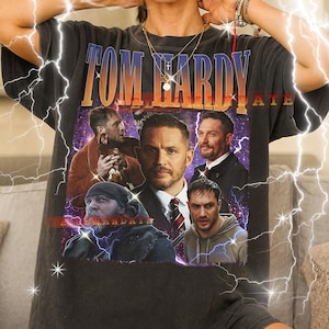 Könnte beinhalten: Dunkelgraues Unisex-T-Shirt mit einer Grafik des Schauspielers Tom Hardy und dem Text "Tom Hardy" in Orange und Blau. Das Shirt hat einen Vintage-Stil mit mehreren Bildern des Schauspielers. Das Shirt ist eine Unisex-Größe.