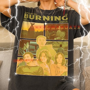 May include: A dark gray t-shirt featuring a movie poster design with the title "BURNING" and images of actors. The shirt has a vintage, worn look. The text "ELOMMANDATE" is also visible.