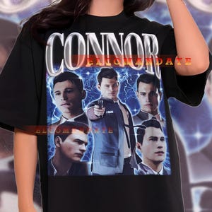 May include: Black t-shirt featuring a graphic of multiple men with the text "CONNOR ENIGMA DATE" in white and orange. The graphic includes the text "ELCOM 800 ANTE". The model is wearing black shorts.