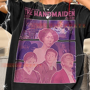Puede incluir: Camiseta negra con un diseño de cartel de película. El gráfico incluye el texto "THE HANDMAIDEN" y varias imágenes de actores. La camiseta tiene cuello redondo y mangas cortas.