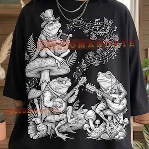 Könnte beinhalten: Schwarzes T-Shirt mit einer Illustration von drei Fröschen, die Musik machen. Ein Frosch spielt Harfe, ein anderer Banjo und der dritte Gitarre. Noten und das Wort "ROMANTE" sind ebenfalls abgebildet.