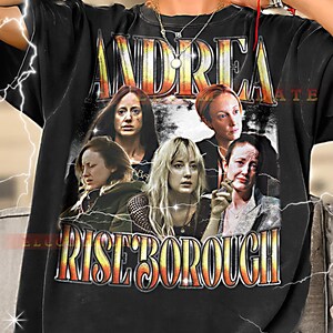 May include: Black t-shirt with a vintage-style graphic featuring the name "ANDREA RISEBOROUGH" and images of several women. The shirt is worn by a person.