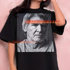 May include: Black t-shirt featuring a grayscale portrait of a man. The text "WELCOME MANDATE" is printed across the image. The model is wearing black shorts and is leaning against a pink wall.