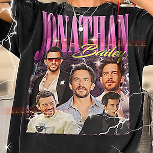 May include: Black t-shirt featuring a collage of images of Jonathan Bailey, with the name "Jonathan Bailey" in large pink letters and "Bailey" in yellow script. The shirt has a vintage aesthetic with lightning bolt graphics.