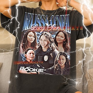 以下が含まれることがあります： 女優メリッサ・オニールと「Lucy Chen」と「The Rookie」の文字がプリントされた、ダークグレーのユニセックスTシャツ。 ヴィンテージスタイルで、かすれたような風合いです。