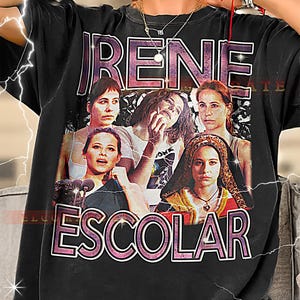 May include: Black t-shirt featuring a graphic design with the name "IRENE ESCOLAR" in a pink and white outlined font. The design includes images of several women. The shirt is a crew neck and appears to be made of cotton.