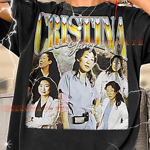 May include: Black t-shirt featuring a graphic of the name "CRISTINA YANG" in gold lettering, with images of a woman in various poses. The shirt has a vintage, distressed design with lightning bolt accents.