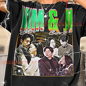 Può includere: T-shirt nera con un design grafico con la scritta "KIM & JI" in verde e oro, insieme a immagini di persone. Il design ha un'estetica vintage. La maglietta è indossata da una persona.