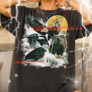 Könnte beinhalten: Dunkelgraues Unisex-T-Shirt mit einer Grafik von Adlern, einem Vollmond und dem Text "BLOOM MANDATE". Das Design enthält rote, weiße und schwarze Elemente. Das Shirt ist in Größe 2XL erhältlich.