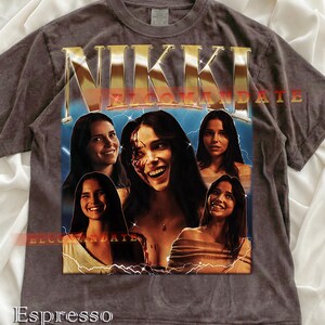 Może przedstawiać: T-shirt w kolorze espresso z grafiką z napisem "NIKKI" w złotych literach. Projekt zawiera wiele zdjęć kobiety z akcentami błyskawic. Widoczny jest r&oacute;wnież tekst "XL COMMANDATE".