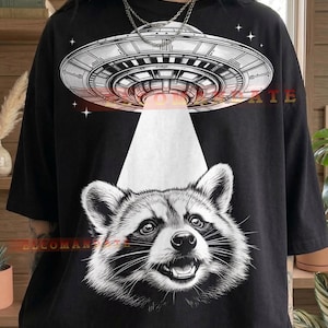 May include: Black t-shirt with a graphic of a UFO shining a white light onto a raccoon. The UFO has red and orange detailing. The word "Black" is printed at the bottom of the shirt.