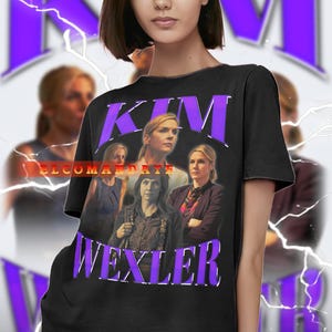 May include: Black t-shirt with a graphic of a woman and the name "KIM WEXLER" in purple text. The shirt has a vintage design with a faded look. The background includes a blurred image and purple lightning bolt graphics.