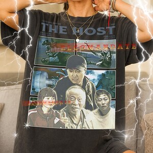 Puede incluir: Camiseta unisex gris oscuro con un diseño de cartel de película que incluye el texto "THE HOST" y varias imágenes. La camiseta tiene un aspecto vintage y desgastado. La modelo lleva un collar y pulseras doradas.