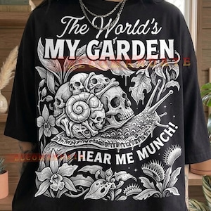 May include: Black t-shirt with a graphic design featuring skulls, a snail, and botanical elements. The text reads "The World's MY GARDEN" and "HEAR ME MUNCH!" in white. The shirt has a relaxed, oversized fit.