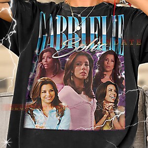 Puede incluir: Camiseta negra con un gráfico del personaje Gabrielle Solis de la serie de televisión. El gráfico incluye múltiples imágenes del personaje con el nombre "Gabrielle Solis" en una fuente estilizada.