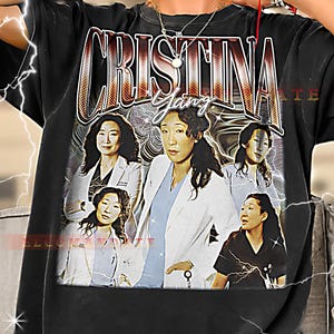May include: Black t-shirt featuring a graphic of the character Cristina Yang from the TV show Grey's Anatomy. The design includes the name "Cristina Yang" in a retro font, along with multiple images of the character.