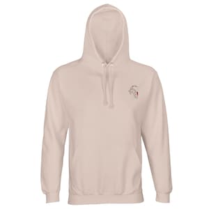 Könnte beinhalten: Ein hellbeige Kapuzenpullover mit Kordelzugkapuze und Kängurutasche. Der Kapuzenpullover hat ein kleines gesticktes Design auf der oberen linken Brust. Das Design ist eine Strichzeichnung einer Ziege mit roten Akzenten.