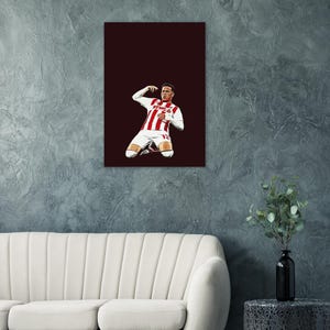 Könnte beinhalten: Gerahmter Kunstdruck mit einem Fußballspieler in einem weiß-rot gestreiften Trikot, der kniet und einen Arm hebt. Der Hintergrund ist ein tiefes Burgunderrot. Das Kunstwerk wird über einem weißen Sofa präsentiert.
