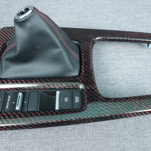 May include: Black carbon fiber car interior trim panel with red accents and a black leather gear shift knob. The panel has buttons labeled "Drive Mode", "Sport", and "Auto Hold".