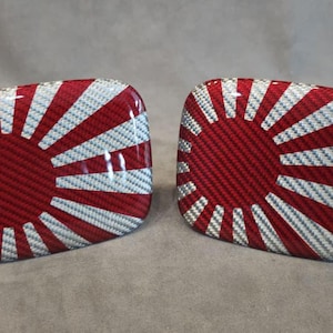 Könnte beinhalten: Zwei passende rote und weiße Carbonfaser-Accessoires, jeweils mit einem Design der aufgehenden Sonne. Die Accessoires haben eine glänzende Oberfläche und ein strukturiertes Carbonfasermuster. Das Design erinnert an die japanische Flagge.