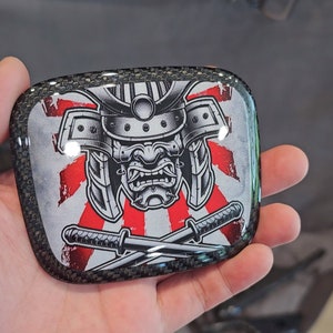 Puede incluir: Una imagen en blanco y negro de un guerrero samurái con un casco con un diseño de sol naciente rojo y blanco detrás de él. Dos espadas se cruzan frente al samurái. La imagen está impresa sobre un fondo de fibra de carbono negro.