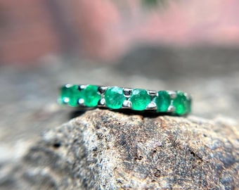Puede incluir: Un anillo de plata con una fila de esmeraldas verdes brillantes. Las piedras preciosas están engastadas juntas, creando una banda continua de color. El anillo se muestra sobre una superficie gris texturizada.
