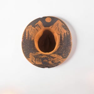 Peut inclure: Un chapeau fedora beige avec une illustration à l'encre noire sur le bord. Le motif représente une chaîne de montagnes, une forêt de pins, une rivière qui coule et un soleil ou une lune circulaire situé au centre supérieur du bord.