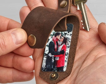 May include: A brown leather keyring with a photo insert featuring a couple in a snowy setting. The keyring has a metal ring and a silver rivet.