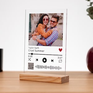 Può includere: Una cornice per foto in acrilico trasparente con una base in legno. La cornice presenta una foto di una coppia, un'interfaccia di lettore musicale con la canzone "Cruel Summer" di Taylor Swift, un'icona a forma di cuore e un codice QR.